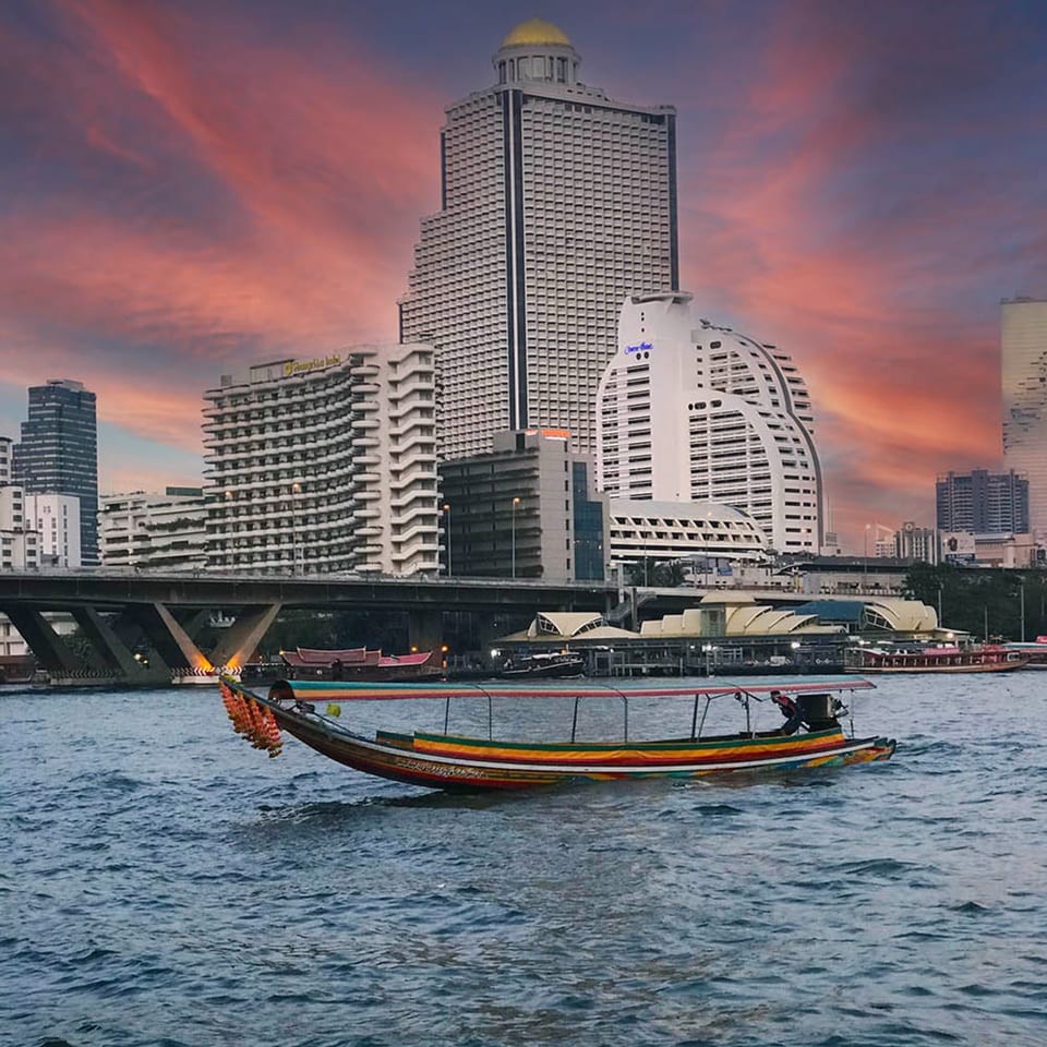 Longtail boat on a river with city buildings and a dramatic sunset sky in the background.