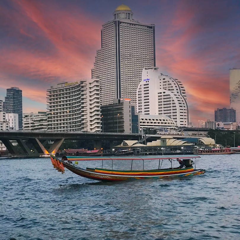 Longtail boat on a river with city buildings and a dramatic sunset sky in the background.