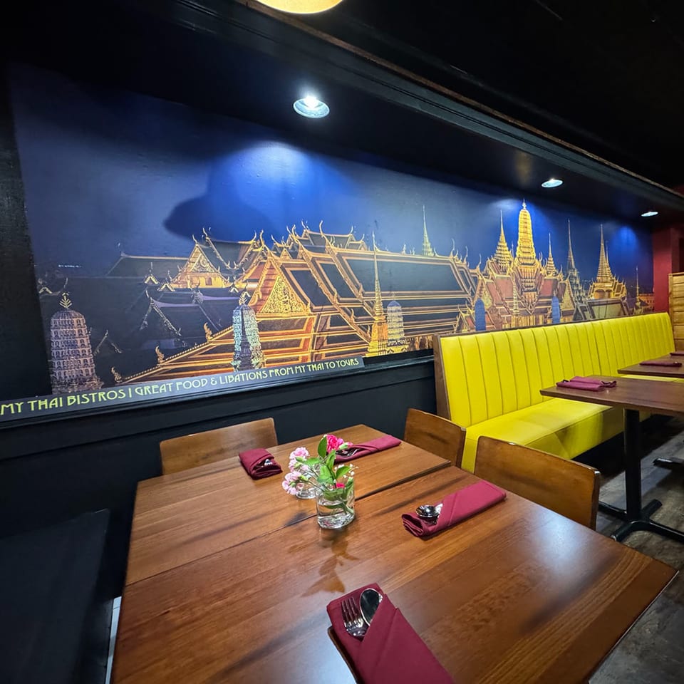 Restaurant interior with wooden tables, red napkins, a yellow booth, and a mural of Thai temples.