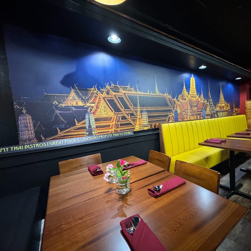 Restaurant interior with wooden tables, red napkins, a yellow booth, and a mural of Thai temples.