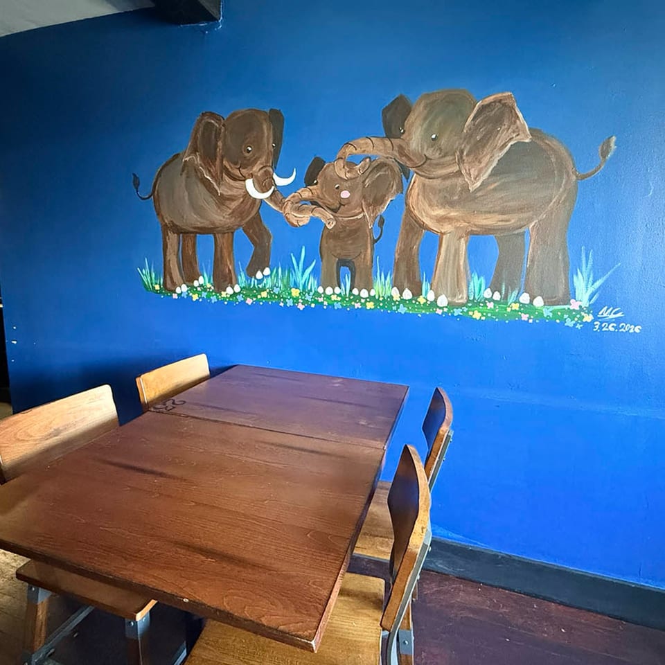 A vibrant blue wall with a painted mural of three elephants, two adults and a baby, above wooden tables.