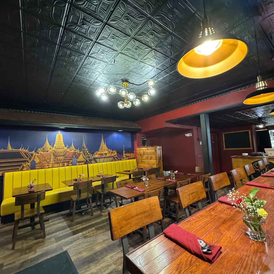 Interior of a Thai restaurant with a mural, yellow booths, wooden tables, and decorative lighting.