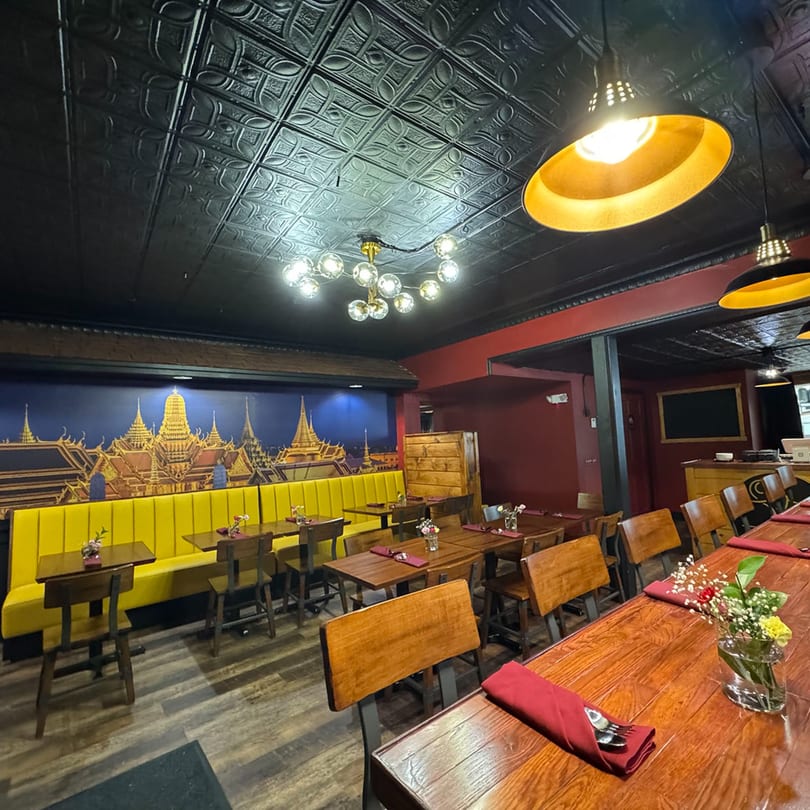 Interior of a Thai restaurant with a mural, yellow booths, wooden tables, and decorative lighting.