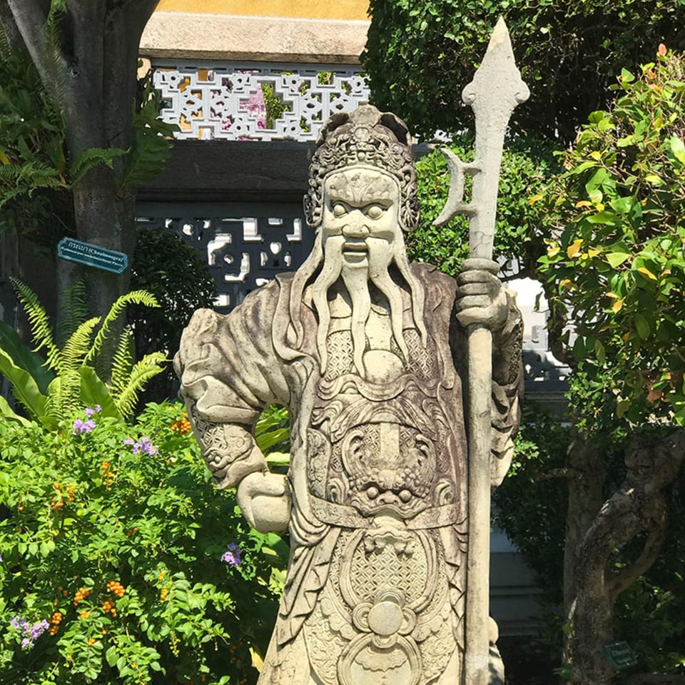 A stone statue of a Chinese guardian with a spear, surrounded by lush green foliage and flowers.