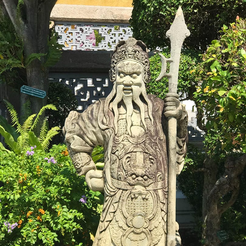 A stone statue of a Chinese guardian with a spear, surrounded by lush green foliage and flowers.
