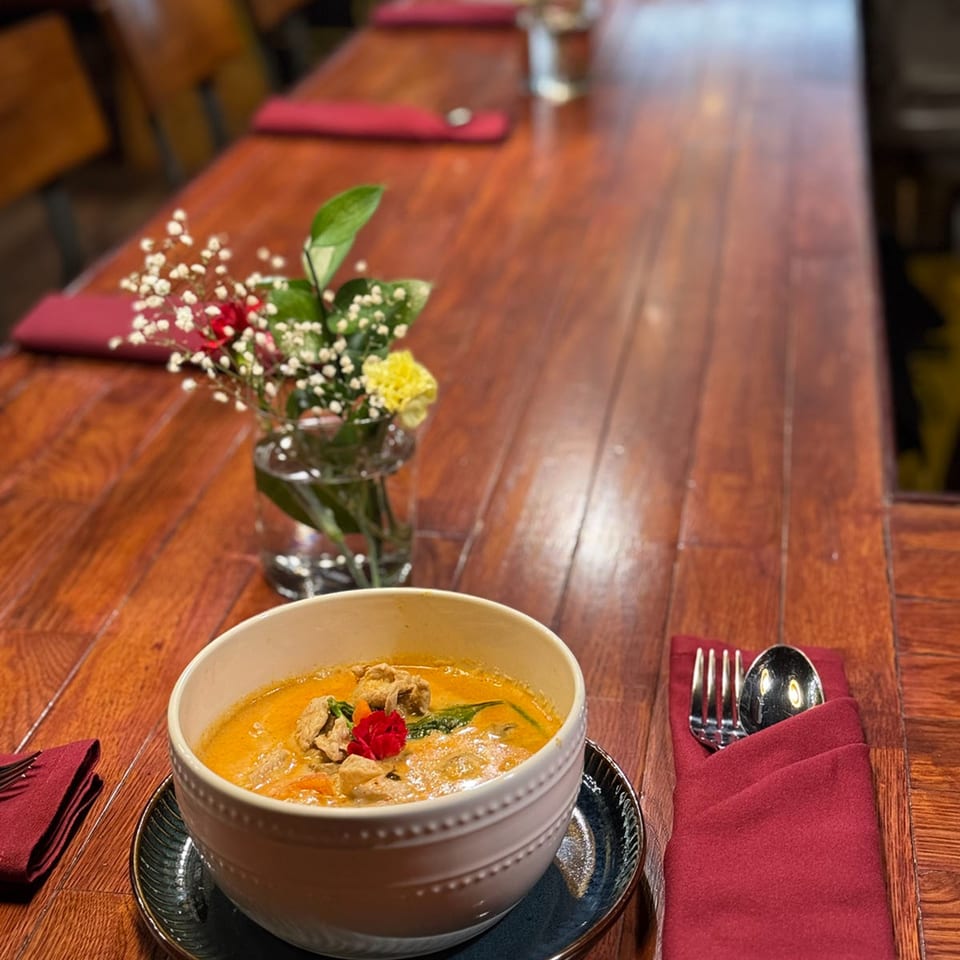 A bowl of curry with chicken and a red flower garnish on a wooden table with a floral centerpiece.