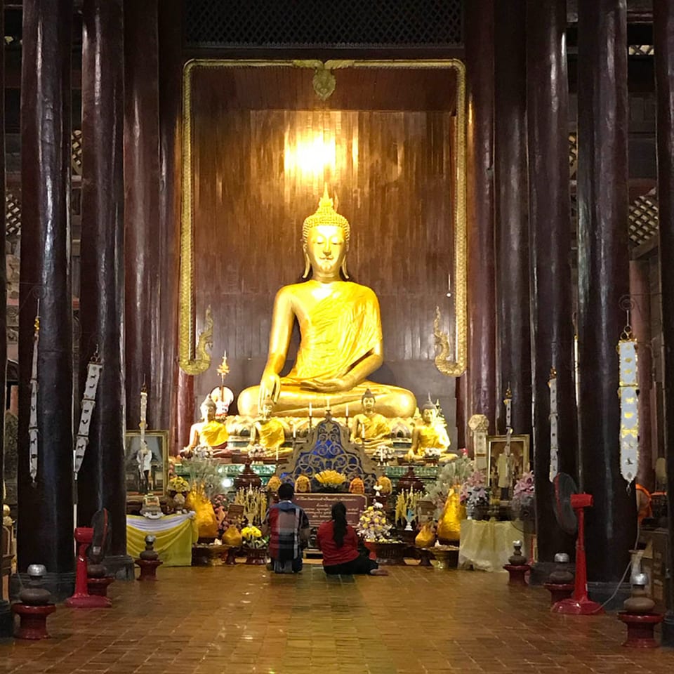 Two people kneeling before a large golden Buddha statue in a dimly lit temple with dark wooden pillars.