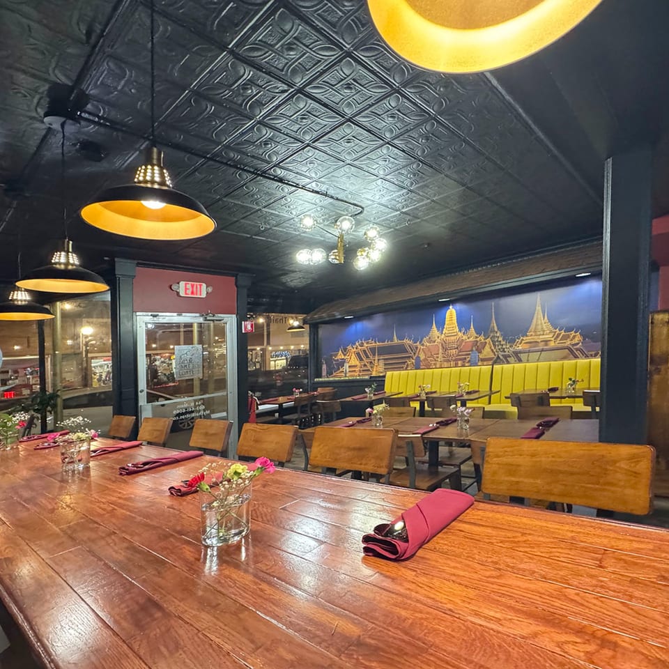 Restaurant interior with a long wooden table, pendant lights, and a mural of Thai temples.