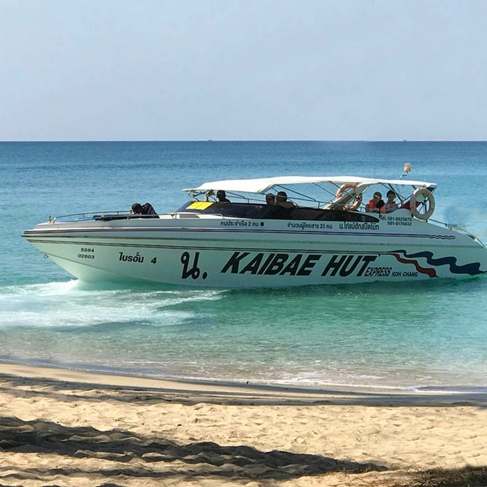 A white speed boat named "KAIBAE HUT EXPRESS" with passengers approaches a sandy beach.
