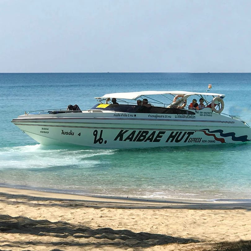 A white speed boat named "KAIBAE HUT EXPRESS" with passengers approaches a sandy beach.