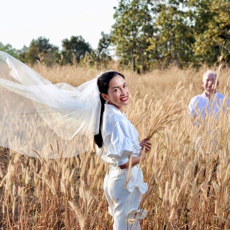 A woman in a white outfit and veil stands in a field of tall grass, holding wheat.