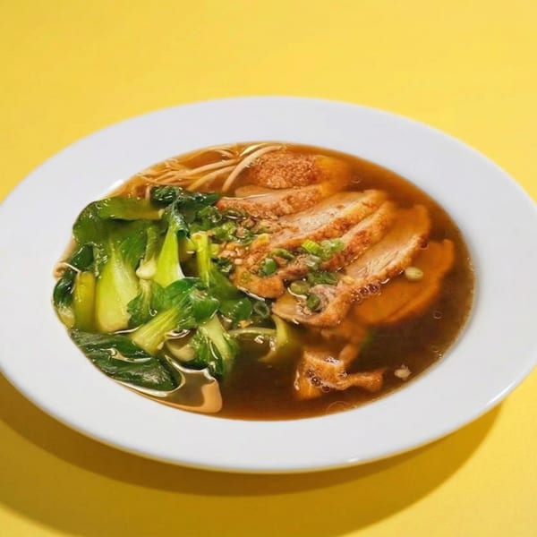 A white bowl of duck noodle soup with bok choy and scallions on a yellow background.