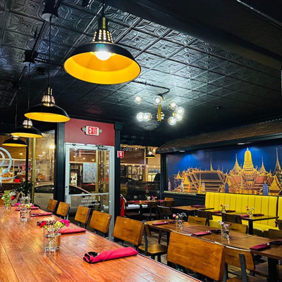 Restaurant interior with long wooden tables, yellow booths, and a mural of a Thai temple.