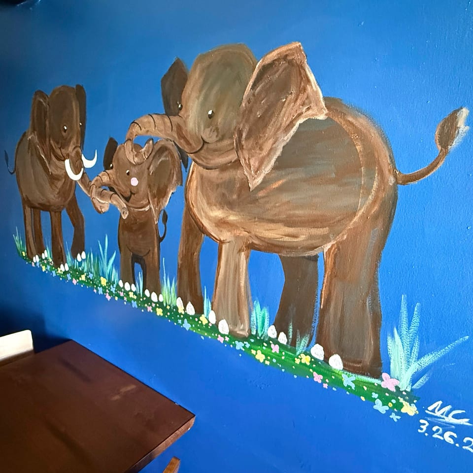 A vibrant blue wall mural depicts a family of brown elephants walking through green grass with colorful flowers.