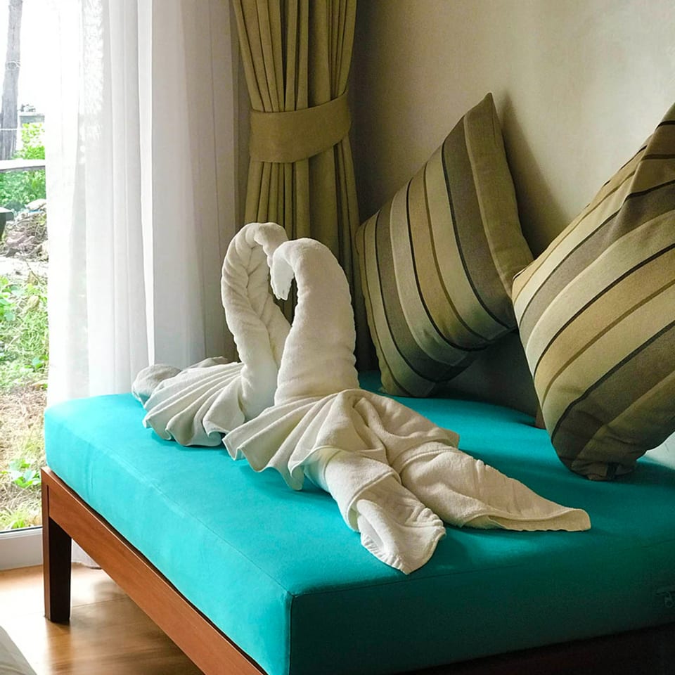 Two white towel swans on a teal daybed with striped pillows by a window with sheer curtains.