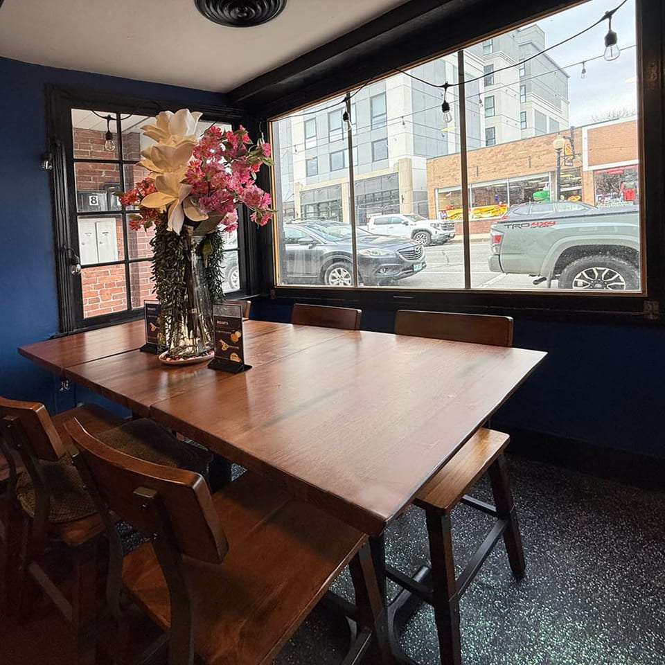 A restaurant interior with a long wooden table, chairs, and a large window overlooking a street.