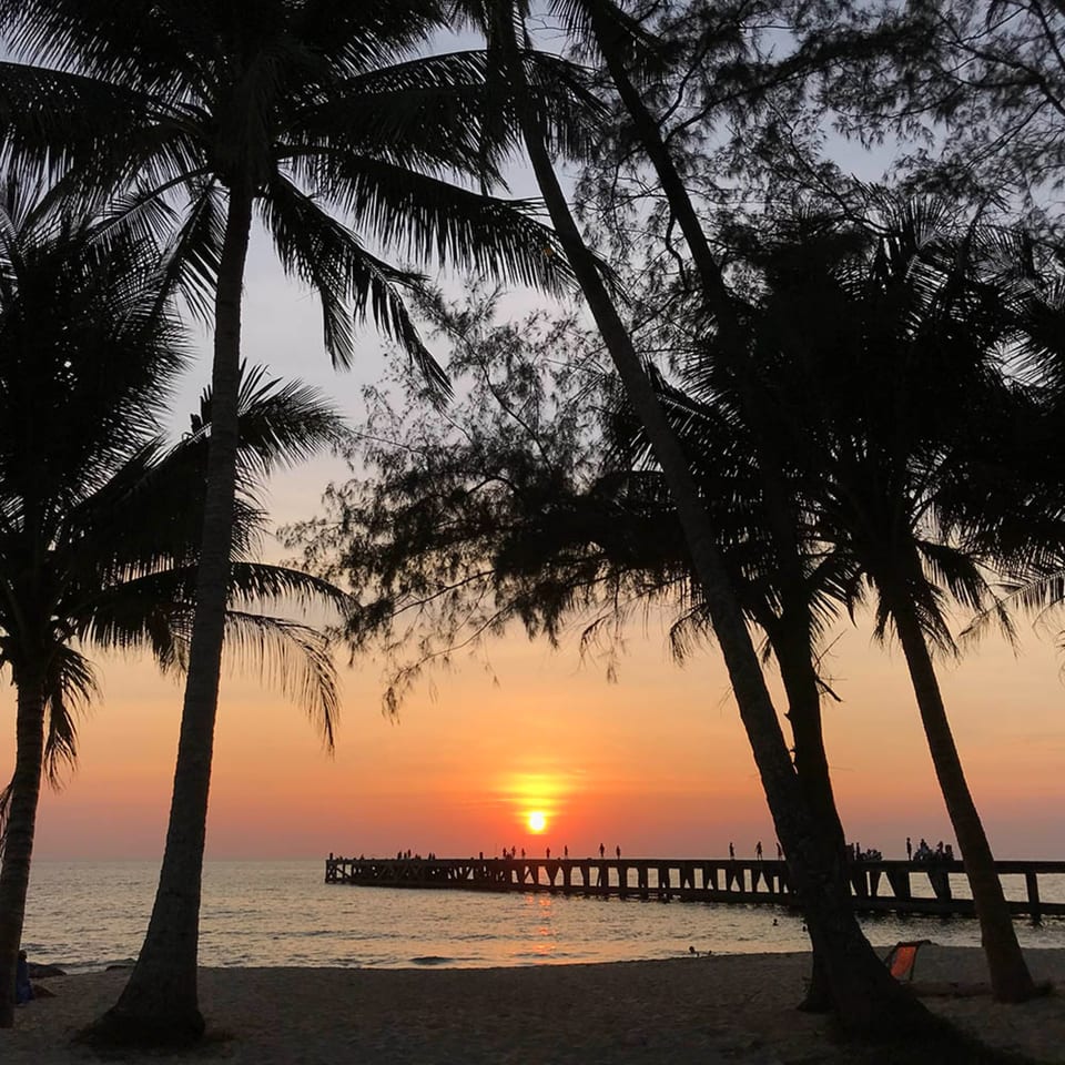 Silhouetted palm trees frame a vibrant sunset over a pier with people on a beach.