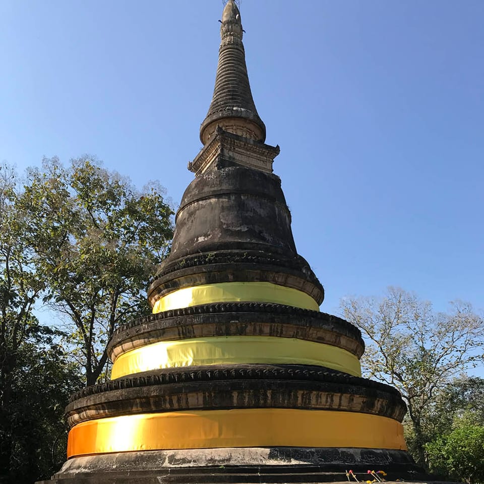 Ancient stone stupa with yellow fabric bands, surrounded by trees under a clear blue sky.