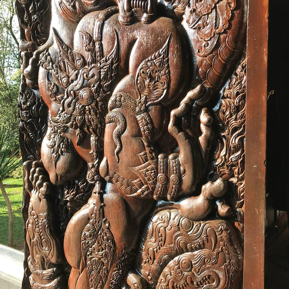 Intricately carved dark wood panel featuring mythical figures and ornate patterns, with greenery in the background.